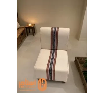 كرسي بتصميم مريح ومبطن بقماش فاخر باللون الأبيض. يتوسط القماش شريط من النقوش التقليدية بالألوان الأحمر والأسود، مما يضفي عليه لمسة جمالية تراثية. للإيجار | استأجر