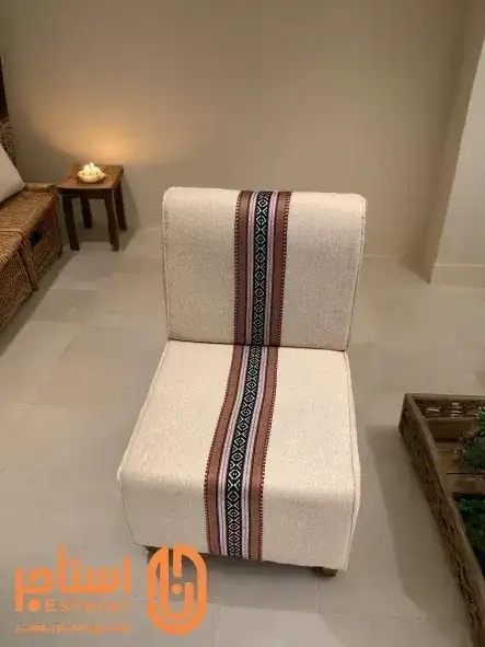 كرسي بتصميم مريح ومبطن بقماش فاخر باللون الأبيض. يتوسط القماش شريط من النقوش التقليدية بالألوان الأحمر والأسود، مما يضفي عليه لمسة جمالية تراثية. - الصورة الرئيسية للإيجار | استأجر