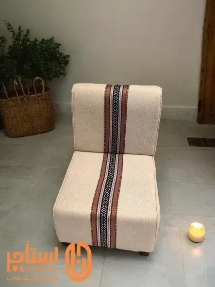 كرسي بتصميم مريح ومبطن بقماش فاخر باللون الأبيض. يتوسط القماش شريط من النقوش التقليدية بالألوان الأحمر والأسود، مما يضفي عليه لمسة جمالية تراثية. - صورة 2 للإيجار | استأجر